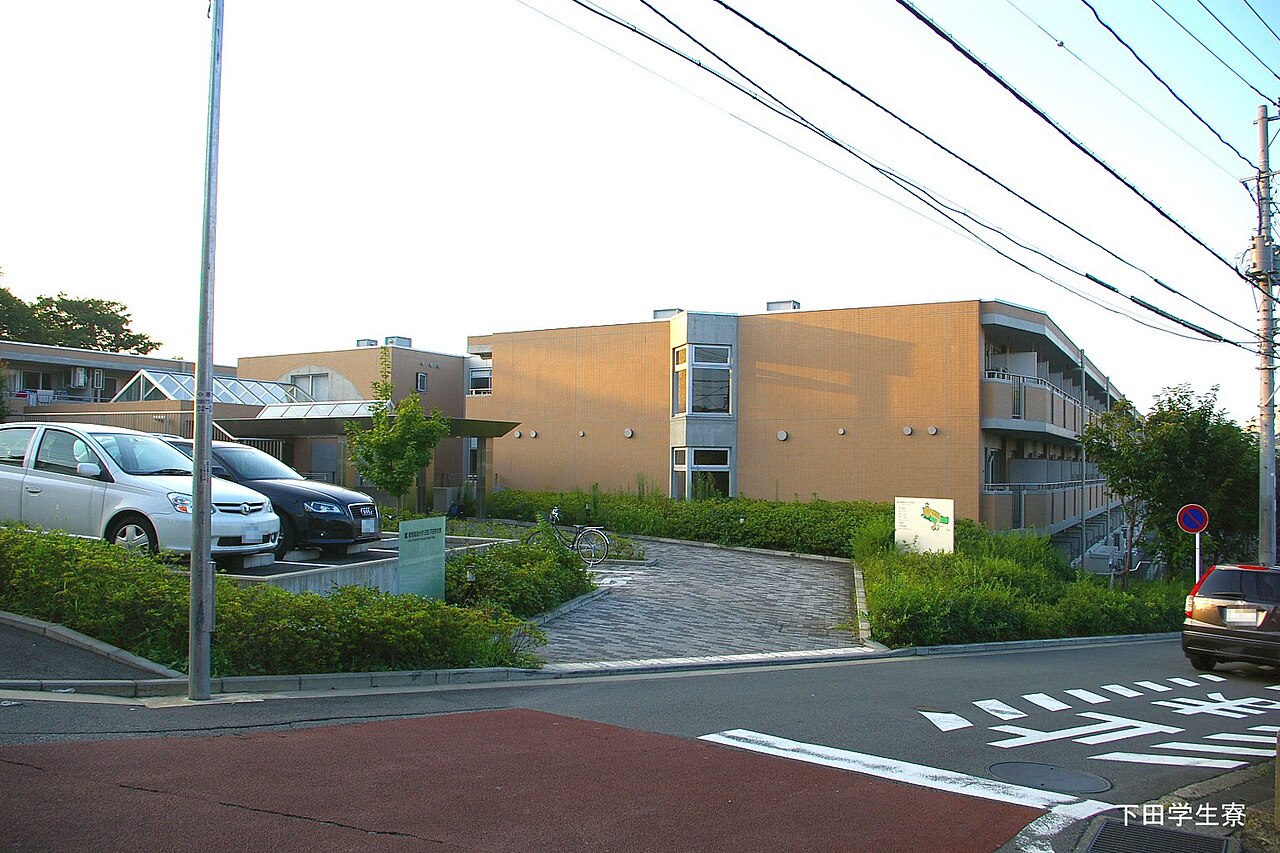 A low university dormitory building behind a car park in a Japanese suburb