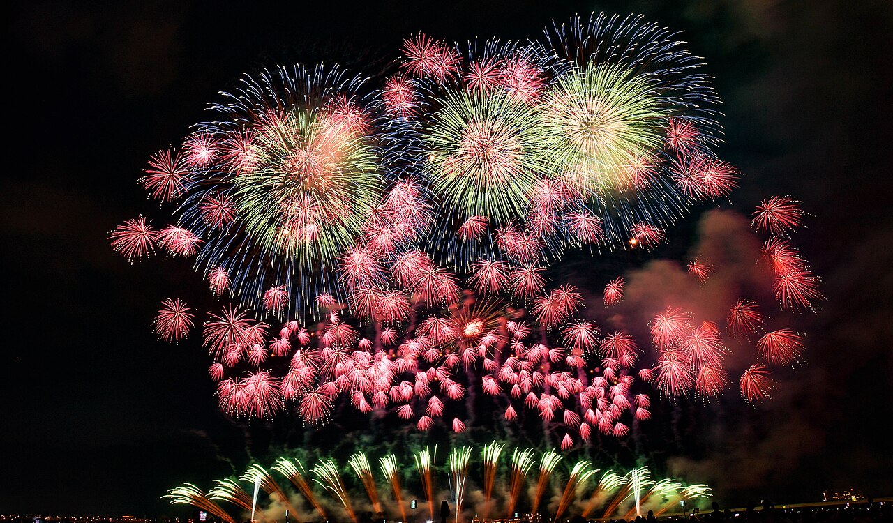A wide spread of large fireworks bursting over a river at the Nagaoka Festival