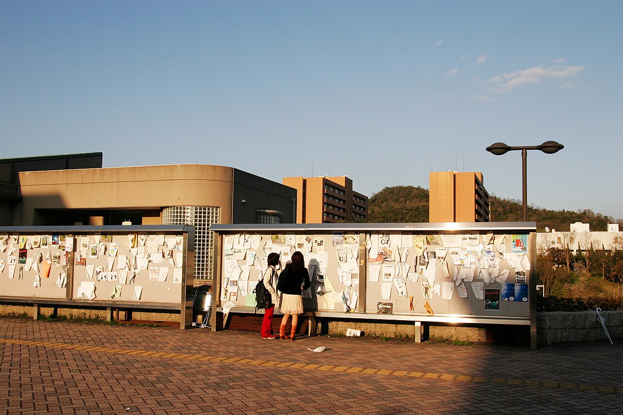 A long outdoor noticeboard on a Japanese university campus, two students reading the notices pinned inside it